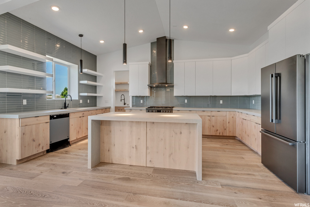 Kitchen featuring wall chimney range hood, appliances with stainless steel finishes, light wood-type flooring, a kitchen island, and tasteful backsplash
