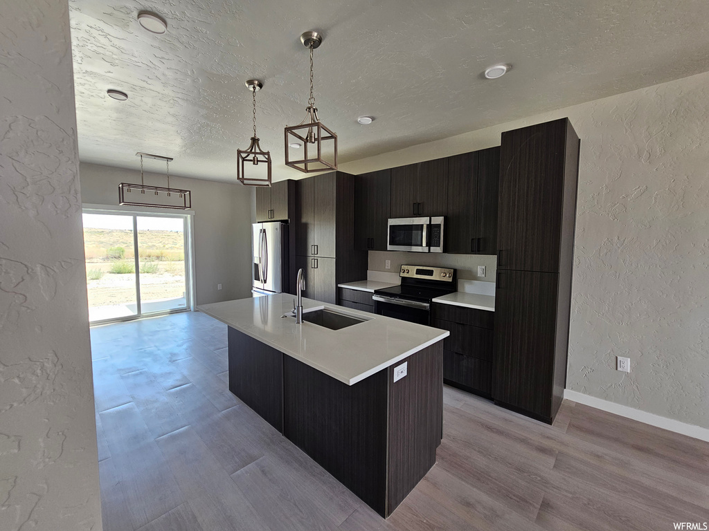 Kitchen featuring dark brown cabinets, stainless steel appliances, a center island with sink, a textured ceiling, light hardwood floors, light countertops, and decorative light fixtures