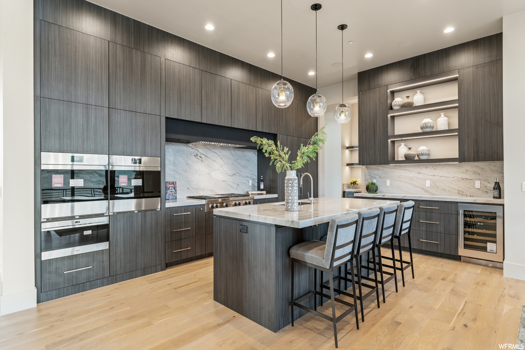 Kitchen featuring decorative light fixtures, kitchen island with sink, backsplash, light hardwood floors, appliances with stainless steel finishes, a center island, dark brown cabinetry, and wall chimney exhaust hood