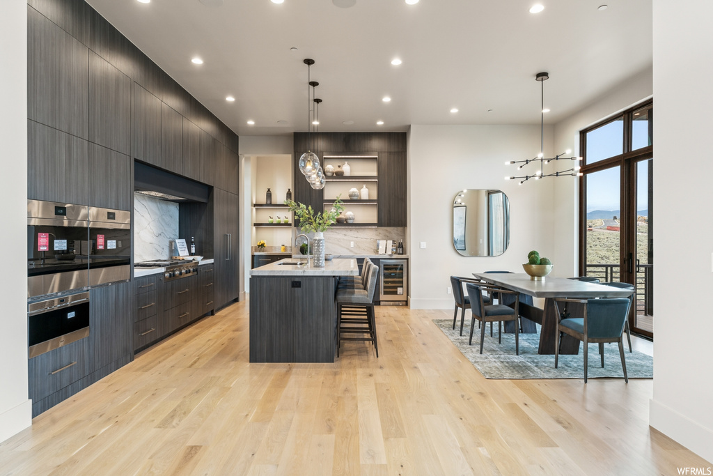 Kitchen featuring hanging light fixtures, an island with sink, backsplash, wall chimney range hood, dark brown cabinets, light countertops, light hardwood floors, a kitchen island, and appliances with stainless steel finishes