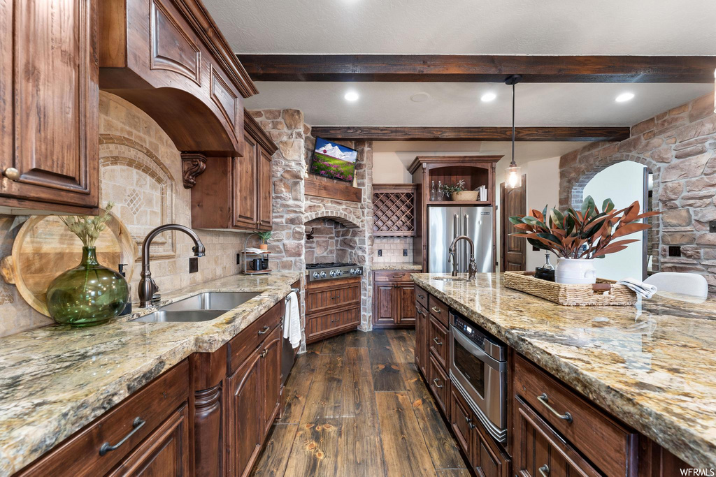 kitchen featuring wood beam ceiling, a fireplace, refrigerator, TV, oven, gas cooktop, kitchen island sink, dark flooring, dark brown cabinetry, light stone countertops, and pendant lighting