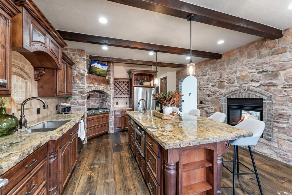 kitchen with wood beam ceiling, a fireplace, a kitchen breakfast bar, gas cooktop, TV, stainless steel refrigerator, kitchen island sink, dark parquet floors, pendant lighting, and light stone countertops