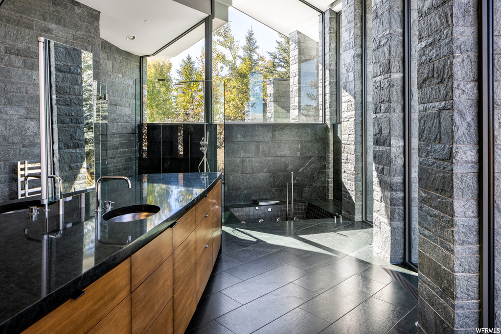 Bathroom with mirror, expansive windows, dark tile flooring, plenty of natural light, vanity, a shower, and tile walls