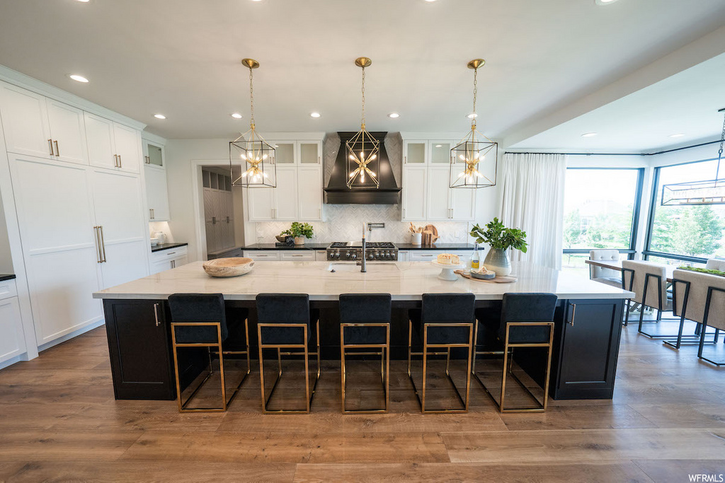 kitchen featuring natural light, a breakfast bar area, exhaust hood, gas stovetop, an island with sink, pendant lighting, light hardwood floors, dark brown cabinetry, and light countertops