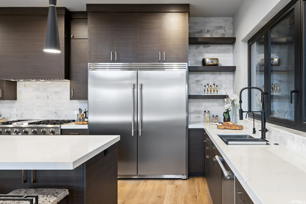 Kitchen with appliances with stainless steel finishes, range hood, dark brown cabinetry, light countertops, backsplash, and light hardwood floors