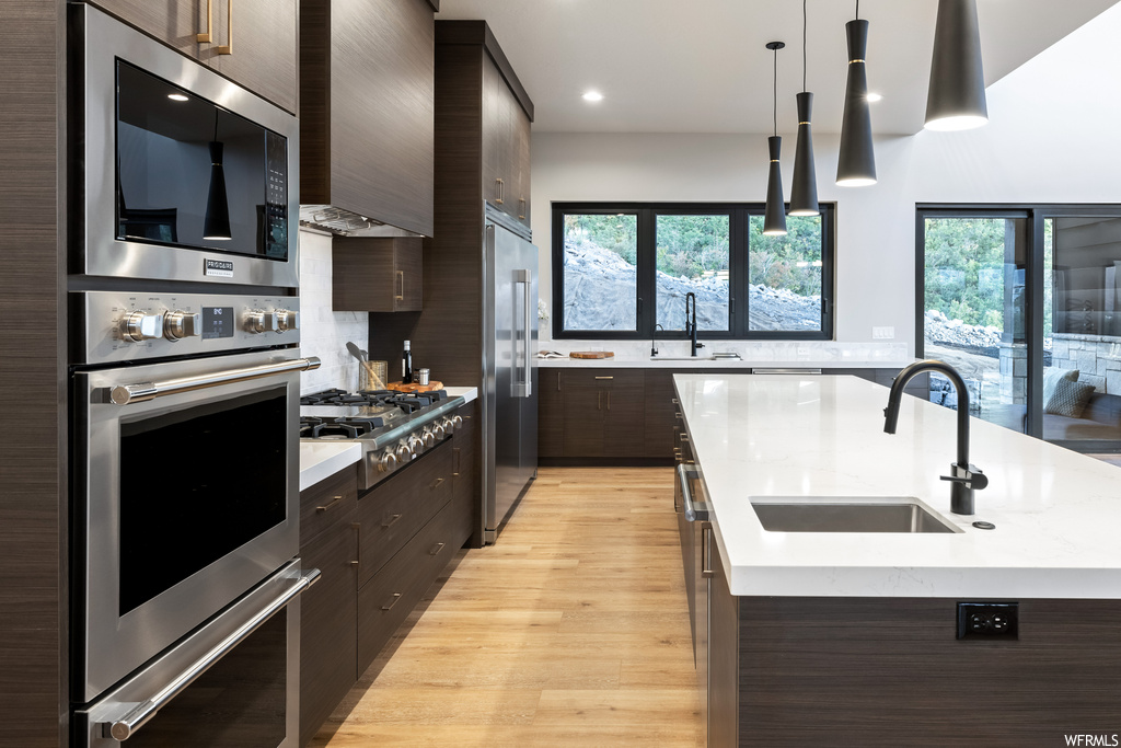 Kitchen featuring wall chimney range hood, decorative light fixtures, built in appliances, light countertops, dark brown cabinets, backsplash, and light hardwood floors