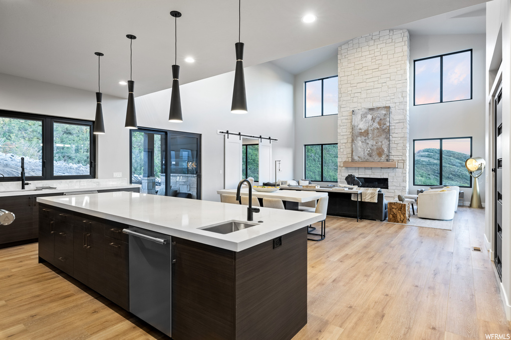 Kitchen with brick wall, stainless steel dishwasher, light hardwood flooring, lofted ceiling, a fireplace, light countertops, dark brown cabinets, hanging light fixtures, kitchen island with sink, and a high ceiling