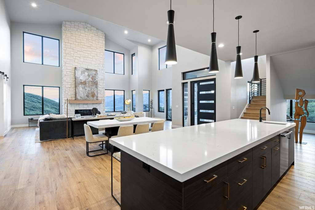 Kitchen featuring stainless steel dishwasher, hanging light fixtures, dark brown cabinets, vaulted ceiling, light hardwood floors, brick wall, light countertops, a center island with sink, a fireplace, and a high ceiling