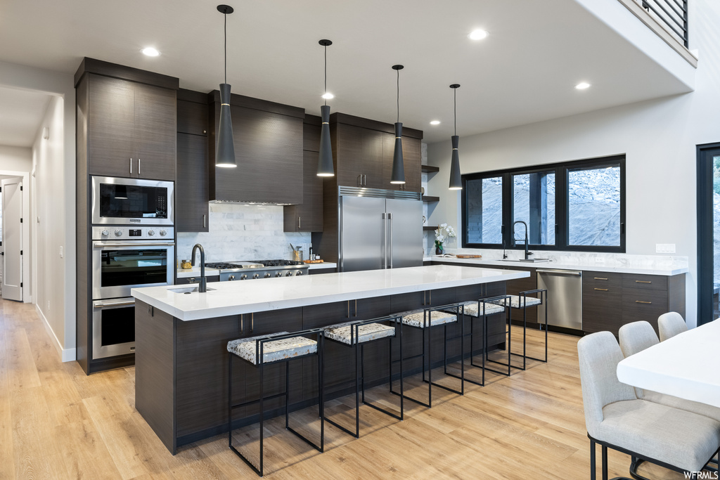 Kitchen with kitchen island with sink, backsplash, built in appliances, dark brown cabinets, light countertops, light hardwood flooring, pendant lighting, and wall chimney exhaust hood