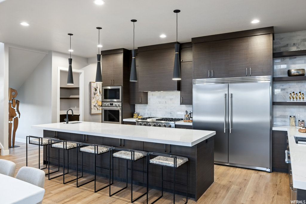 Kitchen with fume extractor, hanging light fixtures, kitchen island with sink, backsplash, built in appliances, dark brown cabinets, light countertops, light hardwood flooring, and stacked washing maching and dryer