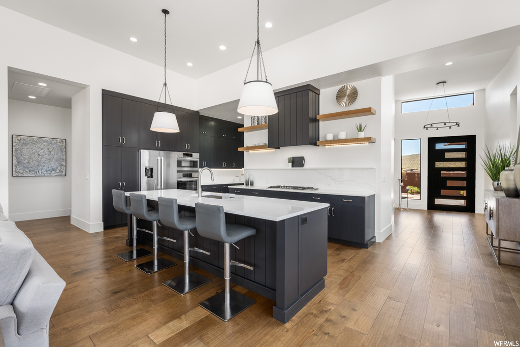 Kitchen with kitchen island with sink, appliances with stainless steel finishes, hanging light fixtures, dark brown cabinetry, light countertops, a high ceiling, and light hardwood floors