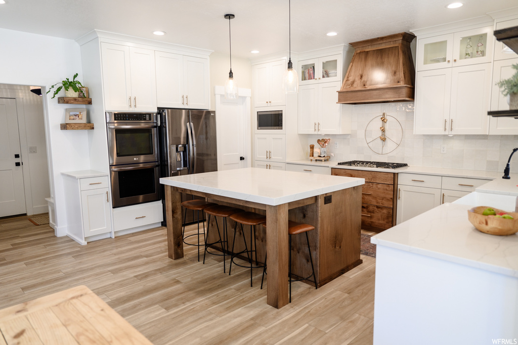 kitchen with a breakfast bar, a kitchen island, gas stovetop, stainless steel double oven, ventilation hood, light hardwood floors, light countertops, pendant lighting, and white cabinetry