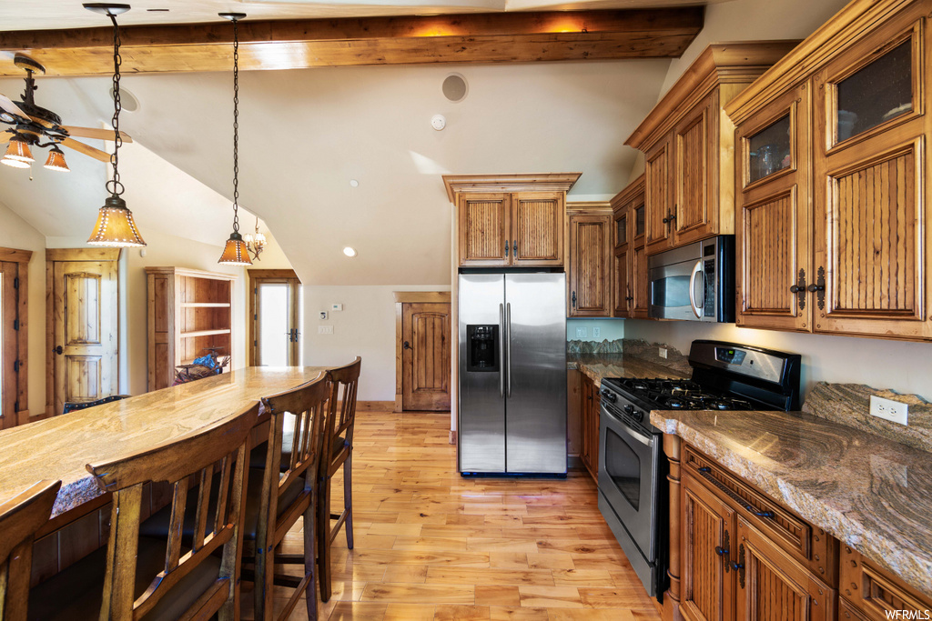 kitchen featuring vaulted ceiling with beams, gas range oven, stainless steel refrigerator, microwave, pendant lighting, brown cabinetry, granite-like countertops, and light hardwood flooring