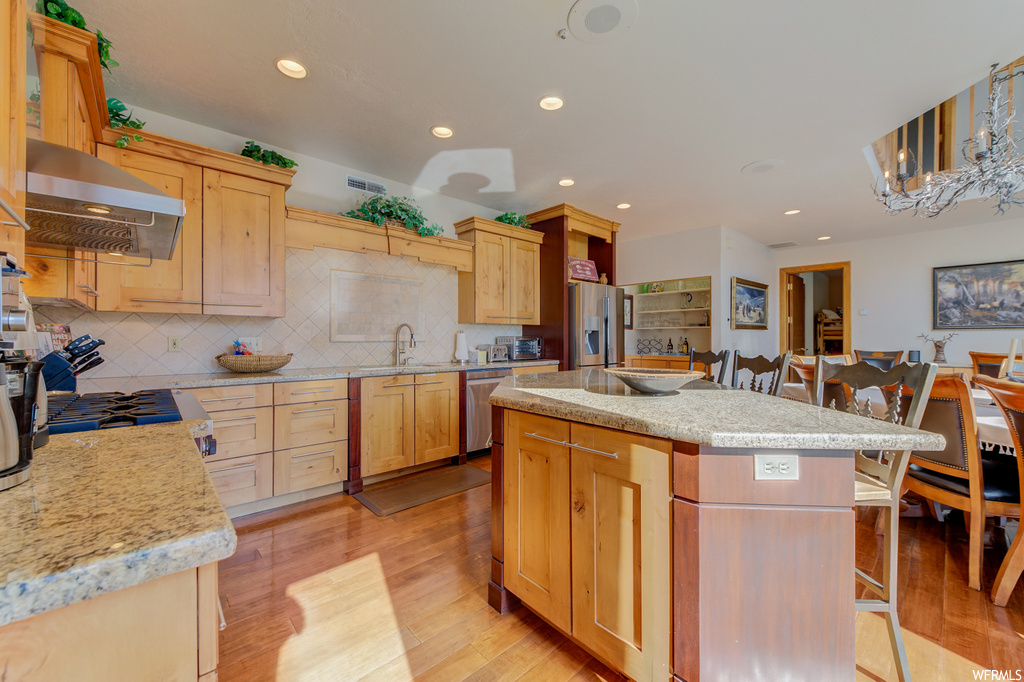 kitchen with a chandelier, stainless steel refrigerator, dishwasher, extractor fan, gas cooktop, light stone countertops, brown cabinetry, and light hardwood flooring