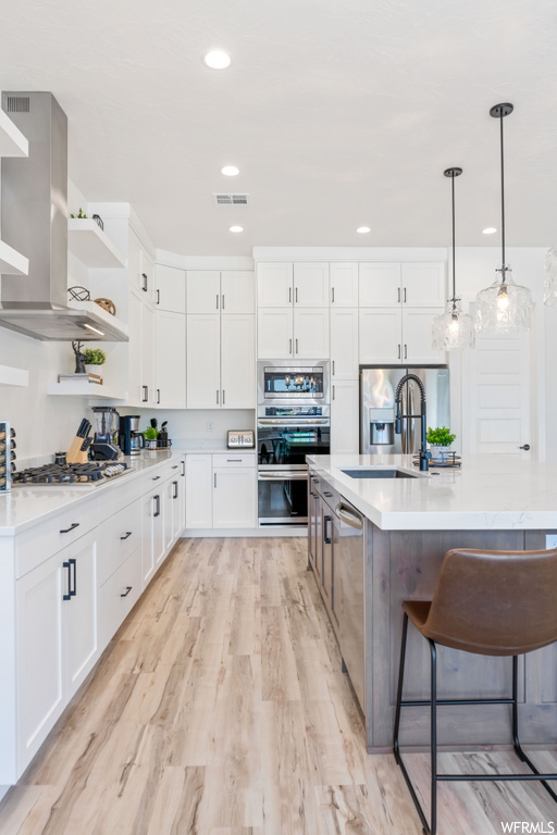 kitchen featuring a kitchen bar, refrigerator, gas stovetop, exhaust hood, oven, dishwasher, light hardwood floors, white cabinets, light countertops, pendant lighting, and kitchen island sink