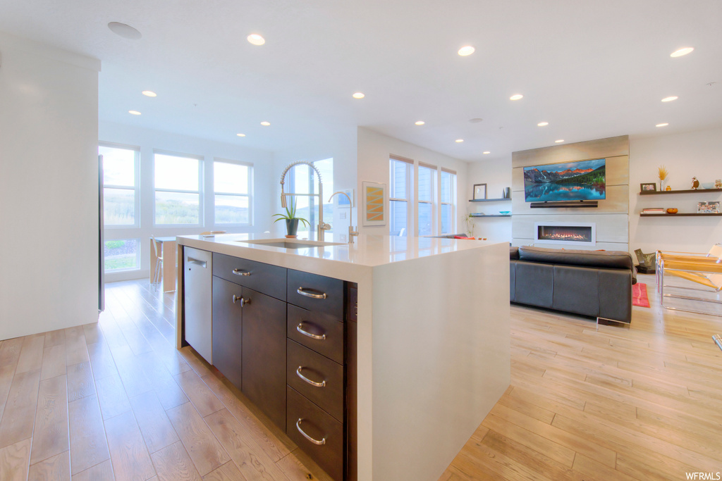 Kitchen with light countertops, white dishwasher, light hardwood flooring, a kitchen island with sink, and a fireplace