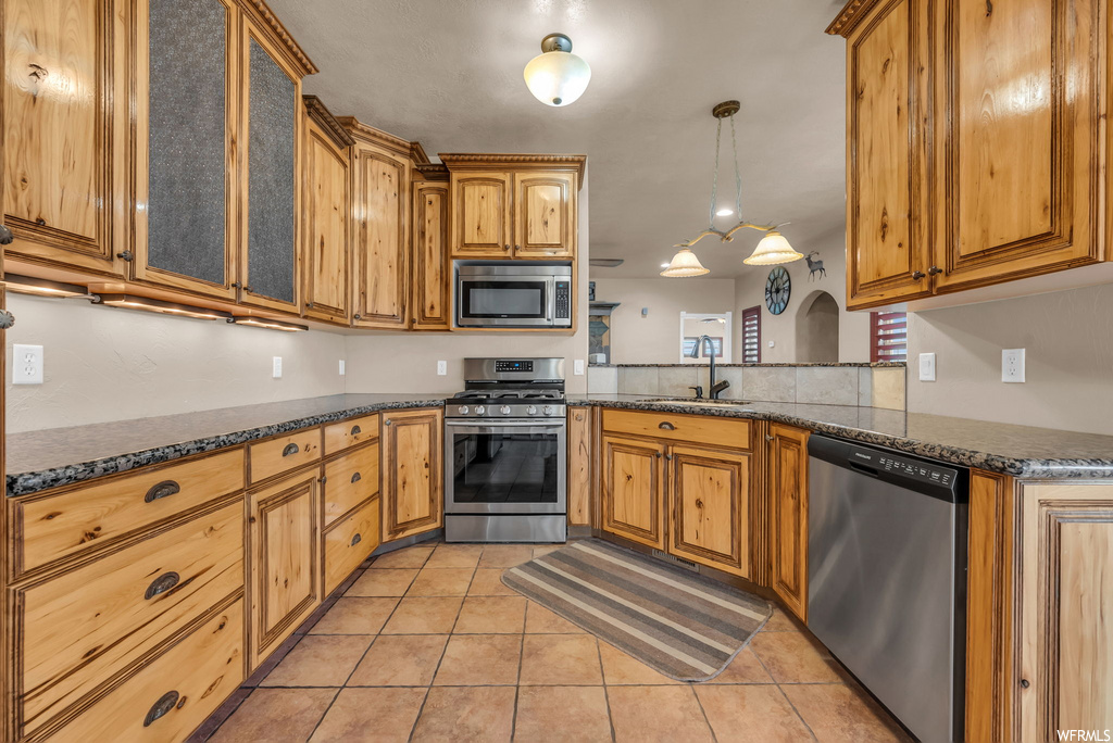 Kitchen with microwave, stainless steel dishwasher, gas range oven, dark stone countertops, light tile flooring, and brown cabinetry