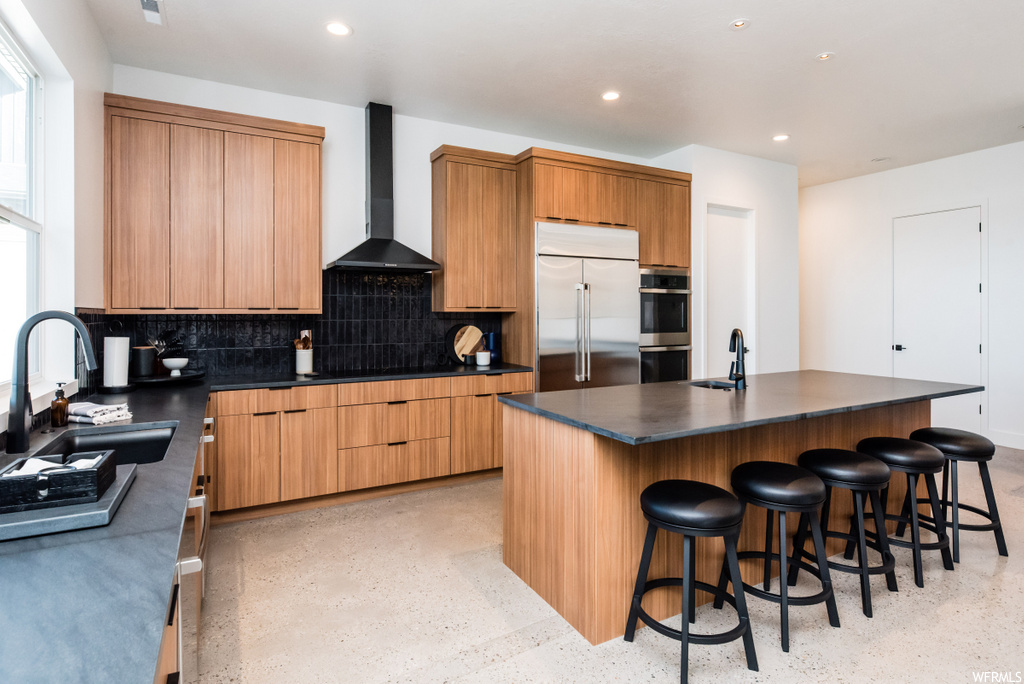 kitchen featuring natural light, a kitchen bar, stainless steel double oven, ventilation hood, refrigerator, dark countertops, brown cabinetry, and light flooring