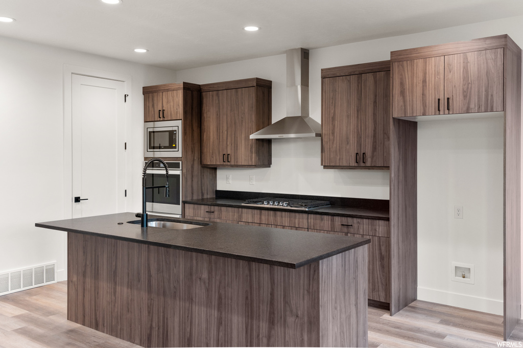 Kitchen with stainless steel appliances, wall chimney range hood, light hardwood flooring, and dark countertops