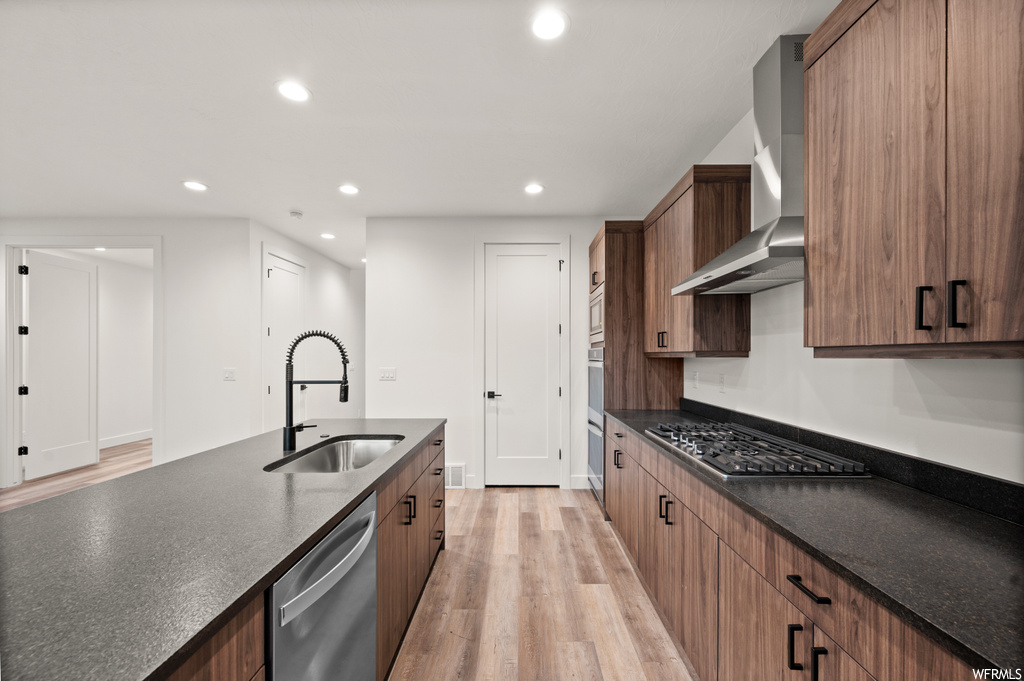 Kitchen featuring dark stone countertops, appliances with stainless steel finishes, wall chimney exhaust hood, and light hardwood floors