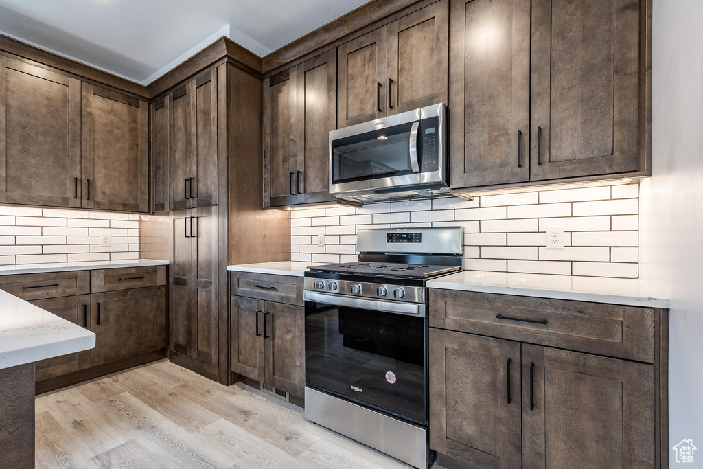Kitchen featuring dark brown cabinetry, tasteful backsplash, light stone countertops, appliances with stainless steel finishes, and light hardwood / wood-style flooring