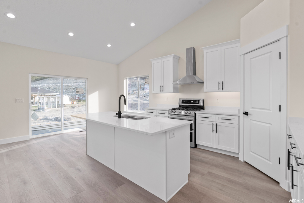 kitchen featuring natural light, vaulted ceiling, range oven, stainless steel finishes, ventilation hood, light countertops, white cabinets, light hardwood floors, and kitchen island sink