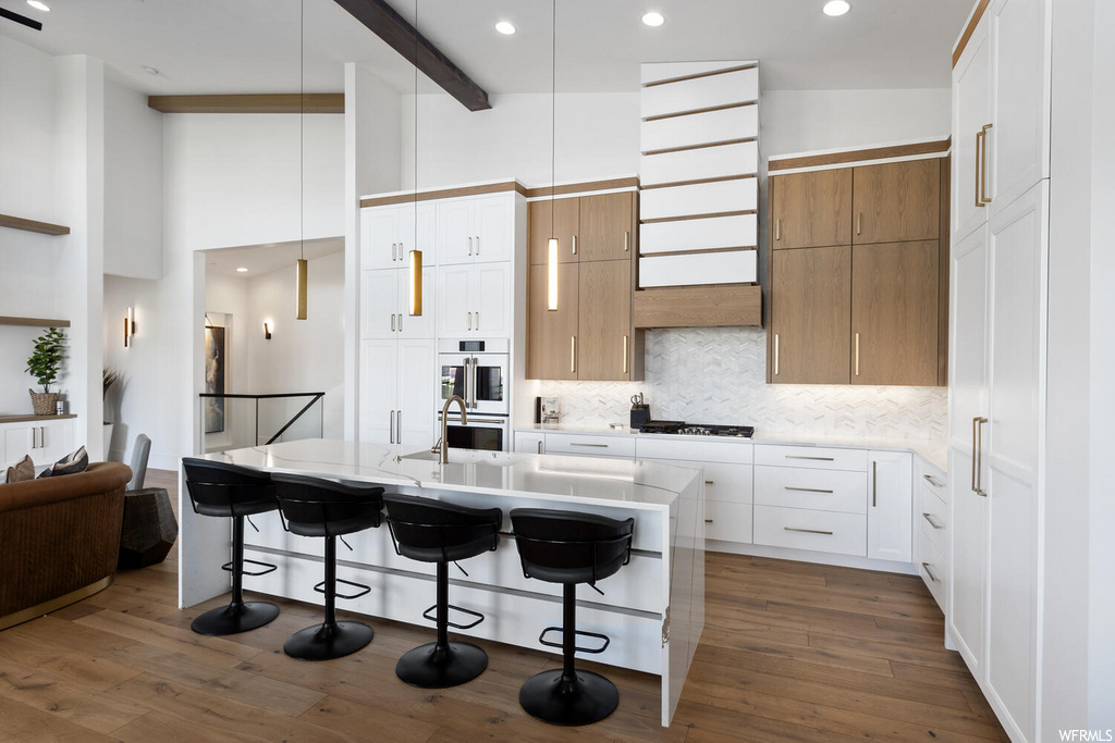 kitchen with hardwood floors, a breakfast bar, stainless steel double oven, gas stovetop, light countertops, white cabinets, kitchen island sink, and pendant lighting