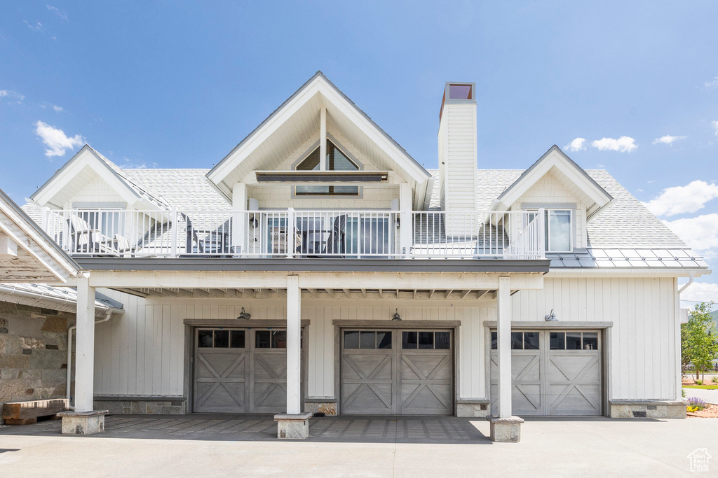 View of front of house with a garage, a balcony, a chimney, driveway, and roof with shingles