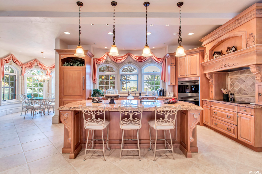 kitchen with a kitchen island, a breakfast bar area, plenty of natural light, electric cooktop, fume extractor, double oven, brown cabinetry, light tile floors, light stone countertops, and pendant lighting