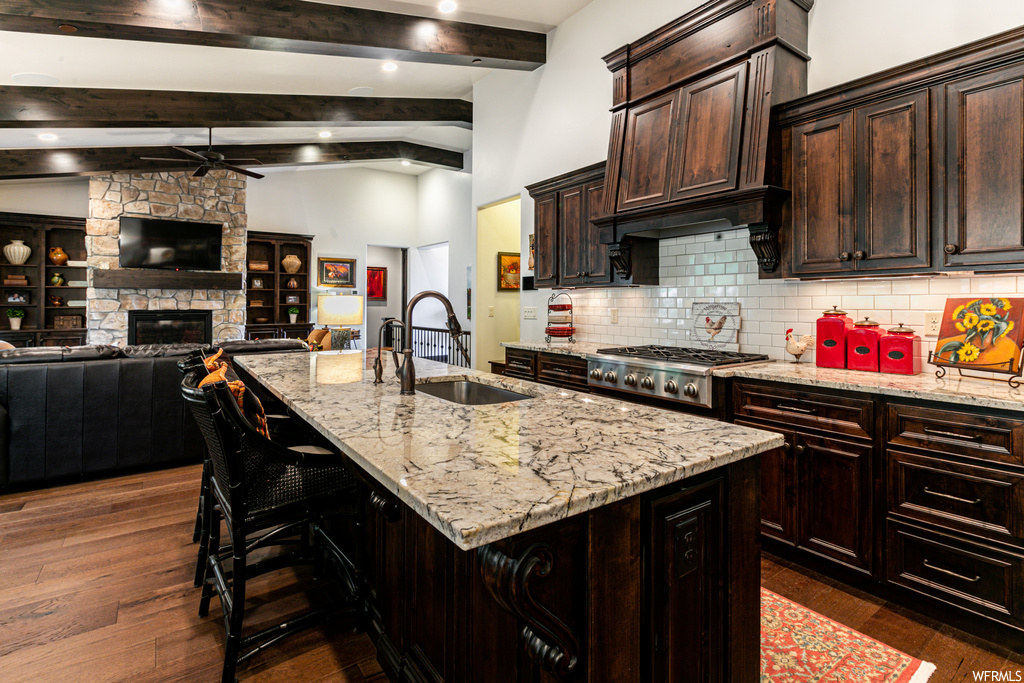 Kitchen featuring wood-type flooring, a fireplace, wood beam ceiling, a kitchen island, TV, gas cooktop, light stone countertops, and dark brown cabinetry