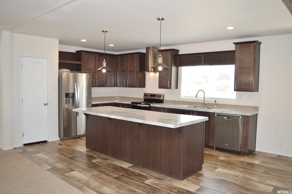 Kitchen featuring wall chimney range hood, light countertops, dark brown cabinets, hanging light fixtures, light hardwood floors, and appliances with stainless steel finishes