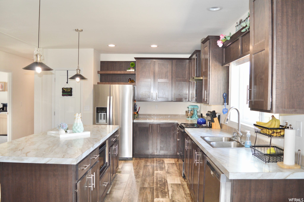 Kitchen with a center island, hardwood floors, refrigerator, stainless steel dishwasher, light countertops, pendant lighting, and dark brown cabinets