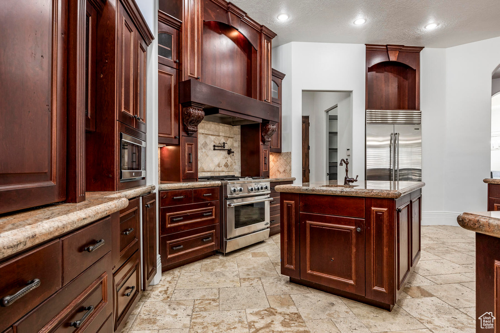 Kitchen with built in appliances, a kitchen island, light tile flooring, wall chimney exhaust hood, and backsplash