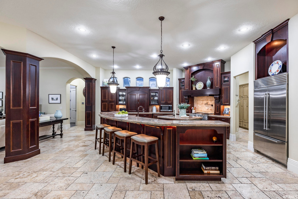 Kitchen featuring a kitchen island with sink, light stone countertops, decorative light fixtures, light tile floors, and stainless steel built in refrigerator
