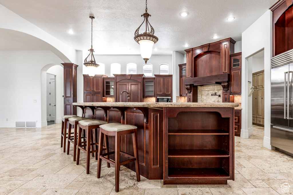 Kitchen with an island with sink, light tile flooring, a kitchen bar, built in fridge, and hanging light fixtures