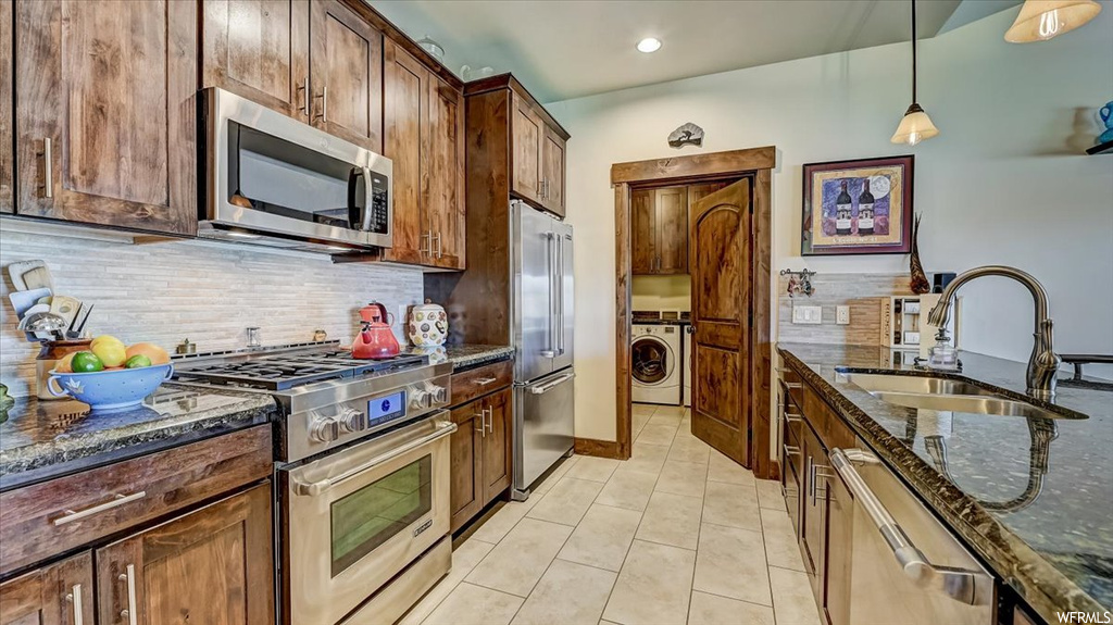 Kitchen featuring gas range oven, stainless steel dishwasher, washer / dryer, refrigerator, microwave, light tile floors, brown cabinetry, dark stone countertops, and pendant lighting