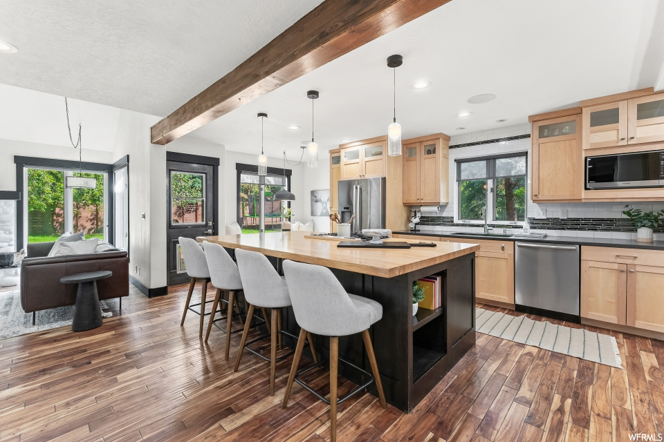 Kitchen featuring a center island, plenty of natural light, a kitchen bar, wood beam ceiling, refrigerator, dishwasher, stainless steel microwave, pendant lighting, light hardwood floors, and light countertops