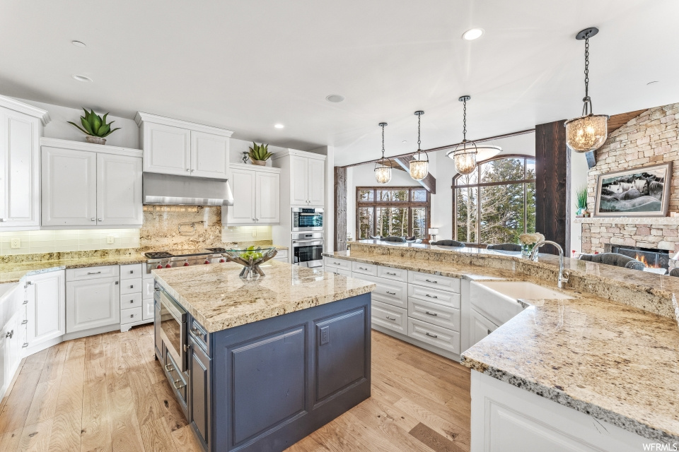 Kitchen with a fireplace, natural light, a kitchen island, exhaust hood, gas stovetop, stainless steel double oven, pendant lighting, white cabinetry, light stone countertops, and light hardwood flooring