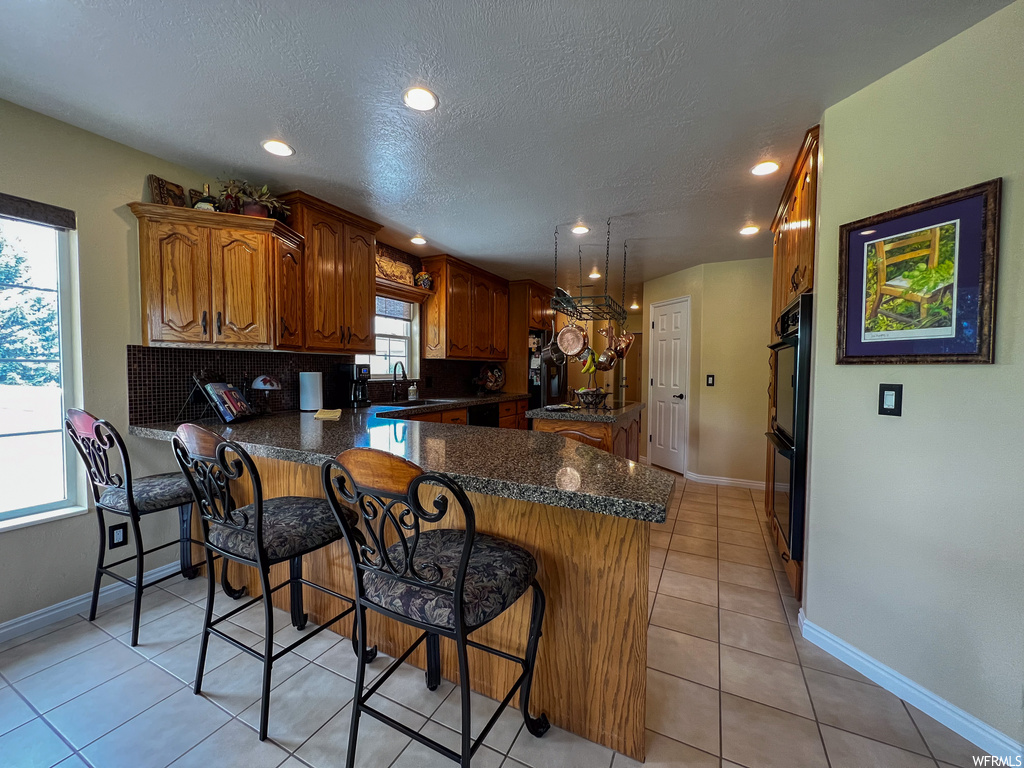 Kitchen featuring natural light, a breakfast bar area, light tile floors, brown cabinets, and dark countertops