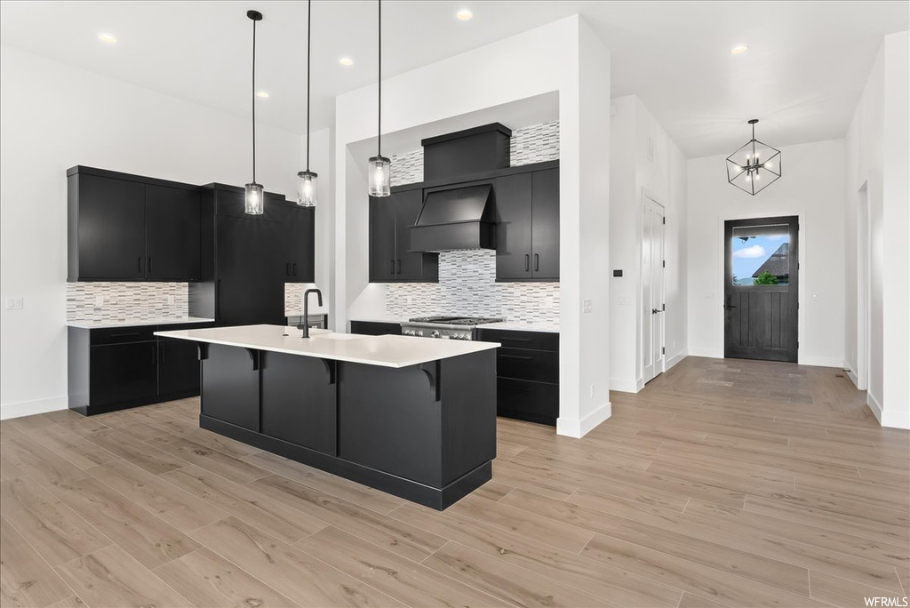 Kitchen with range hood, gas cooktop, stainless steel finishes, kitchen island sink, dark brown cabinets, light hardwood flooring, pendant lighting, and light countertops