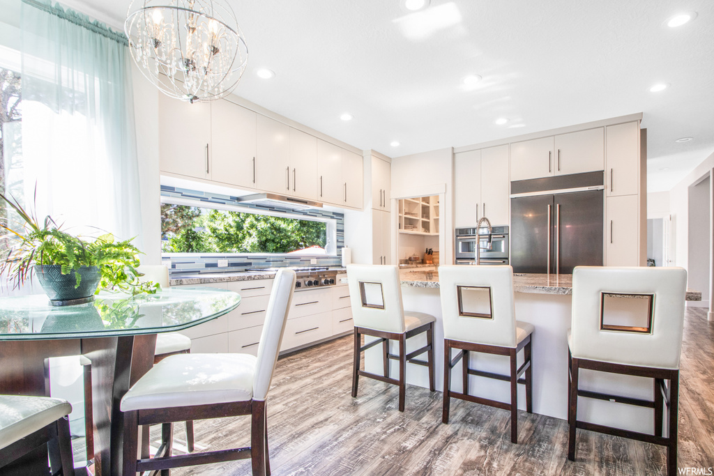 Kitchen with a chandelier, a wealth of natural light, a breakfast bar area, refrigerator, oven, light hardwood floors, and light stone countertops