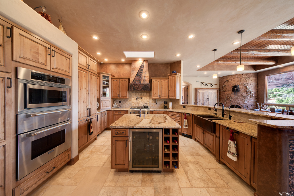Kitchen with skylight, natural light, gas cooktop, stainless steel double oven, extractor fan, brown cabinetry, pendant lighting, light stone countertops, a kitchen island with sink, and light tile floors