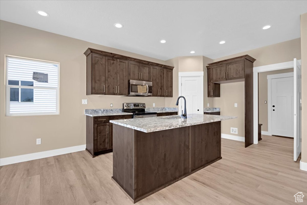 Kitchen with dark brown cabinets, light stone countertops, appliances with stainless steel finishes, light hardwood / wood-style flooring, and a center island with sink