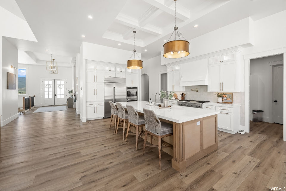 Kitchen featuring coffered ceiling, light countertops, backsplash, custom exhaust hood, light hardwood flooring, beamed ceiling, kitchen island with sink, white cabinetry, hanging light fixtures, and stainless steel appliances