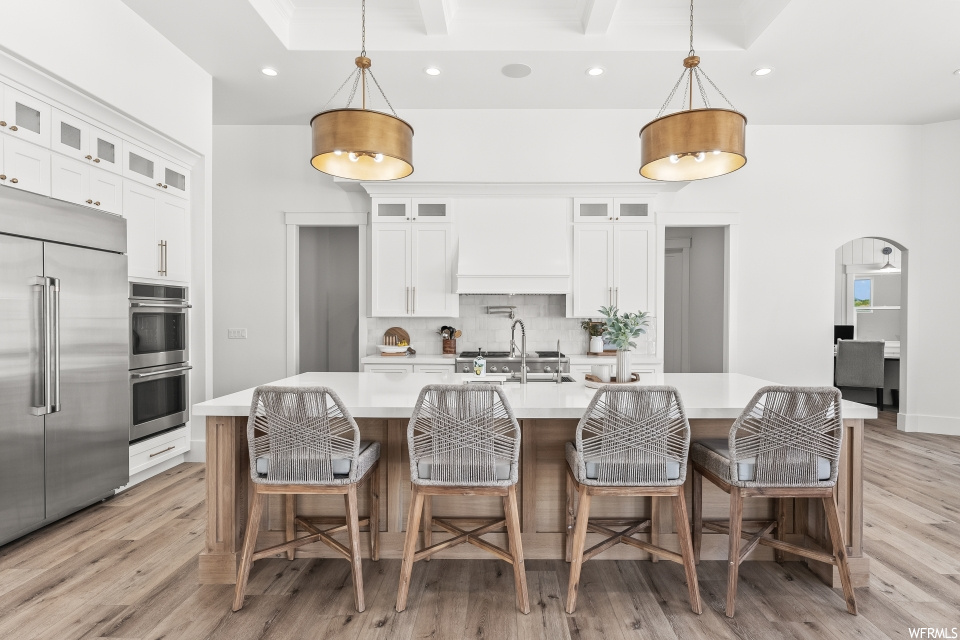 Kitchen featuring a center island, stainless steel appliances including a double oven, light hardwood floors, white cabinetry, light countertops, and pendant lighting