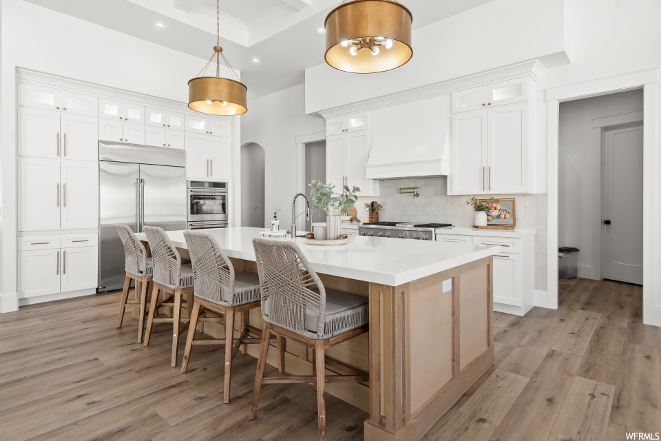 Kitchen with white cabinetry, backsplash, kitchen island with sink, pendant lighting, light countertops, light hardwood flooring, appliances with stainless steel finishes, a center island, and premium range hood