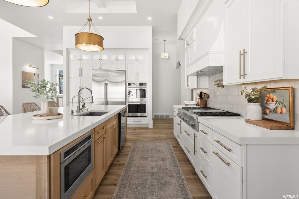 Kitchen with white cabinetry, backsplash, pendant lighting, a raised ceiling, light countertops, wood-type flooring, and stainless steel appliances