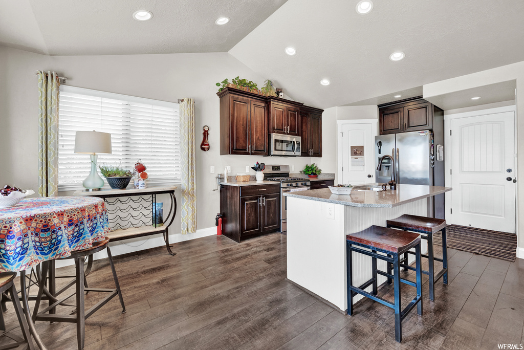 Kitchen with vaulted ceiling, a center island, a kitchen bar, natural light, gas range oven, microwave, refrigerator, light countertops, dark brown cabinetry, and dark parquet floors
