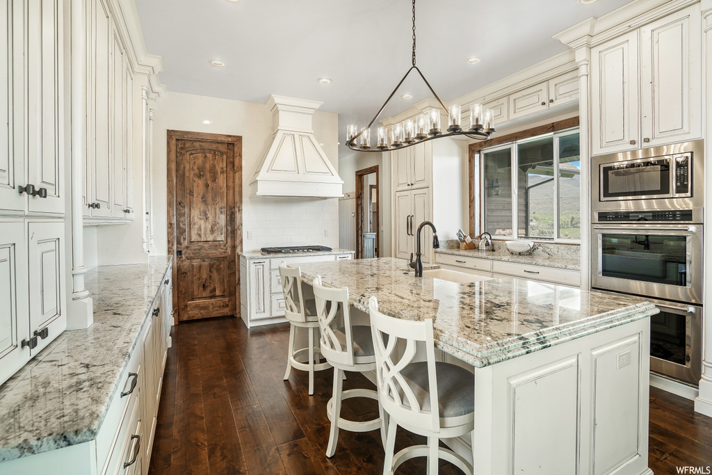 Kitchen featuring a breakfast bar area, stainless steel microwave, range hood, double oven, white cabinets, pendant lighting, light stone countertops, a center island with sink, and dark hardwood floors