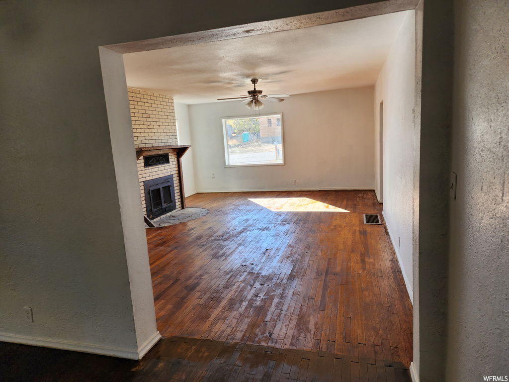 Unfurnished living room featuring dark hardwood / wood-style floors, a fireplace, brick wall, and ceiling fan